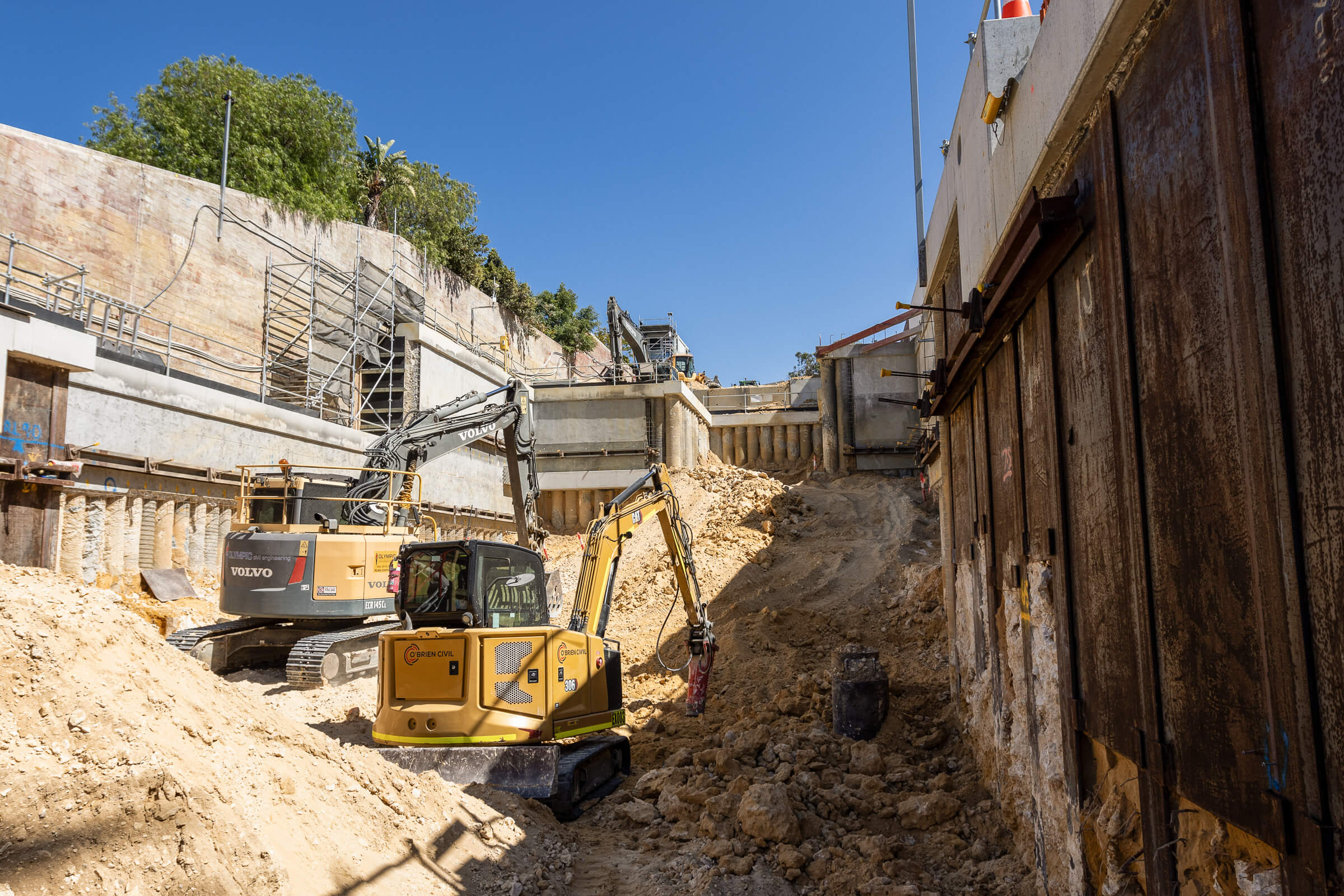 Construction site with two excavators working on a steep dirt slope beside a retaining wall under clear blue sky.