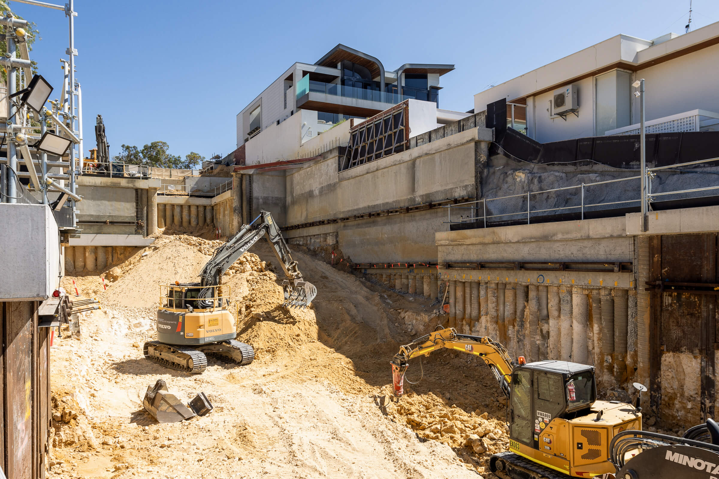 Excavation site with two excavators working in a deep trench surrounded by retaining walls and modern buildings above.