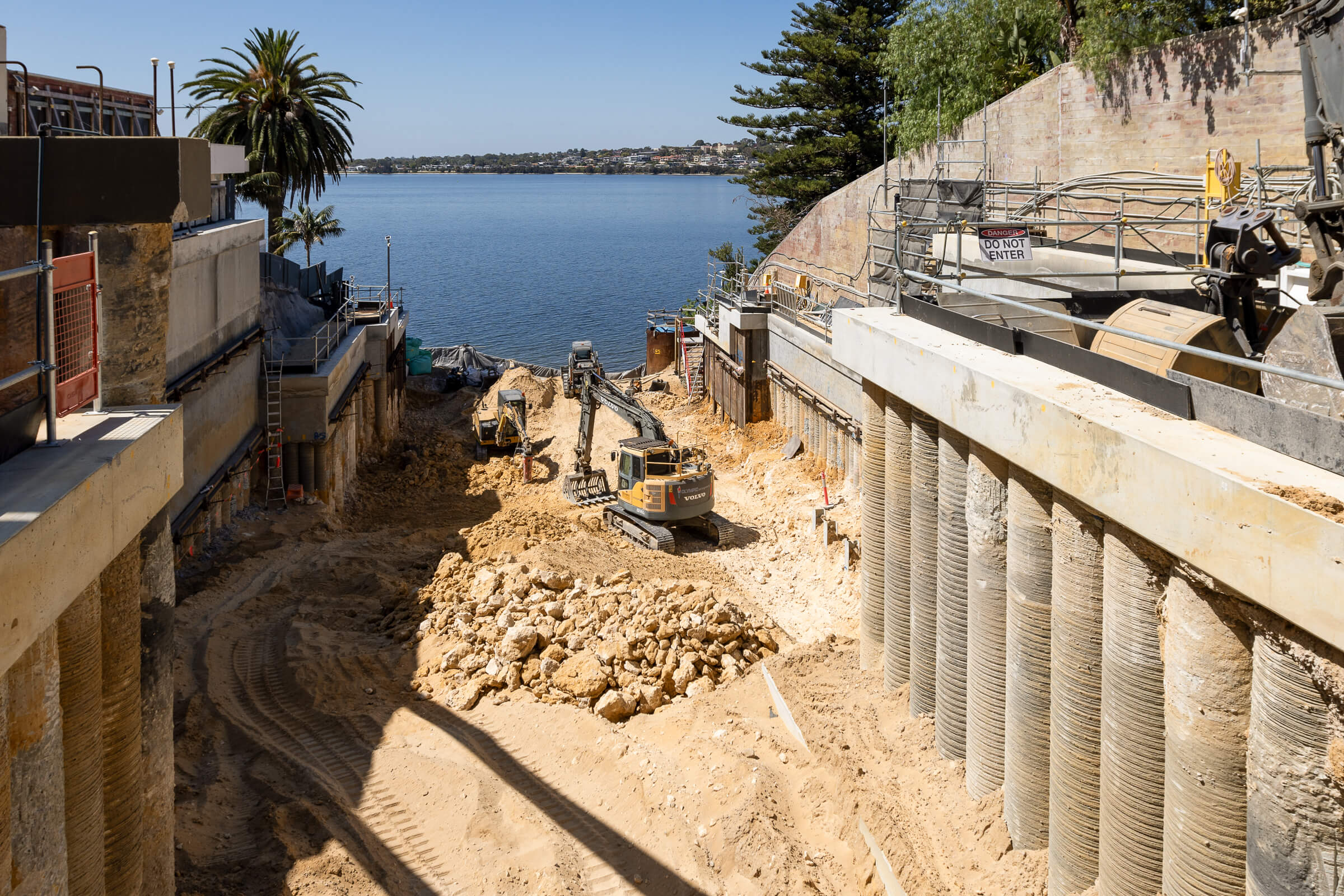Construction site with excavators digging near a body of water, supported by concrete column walls.