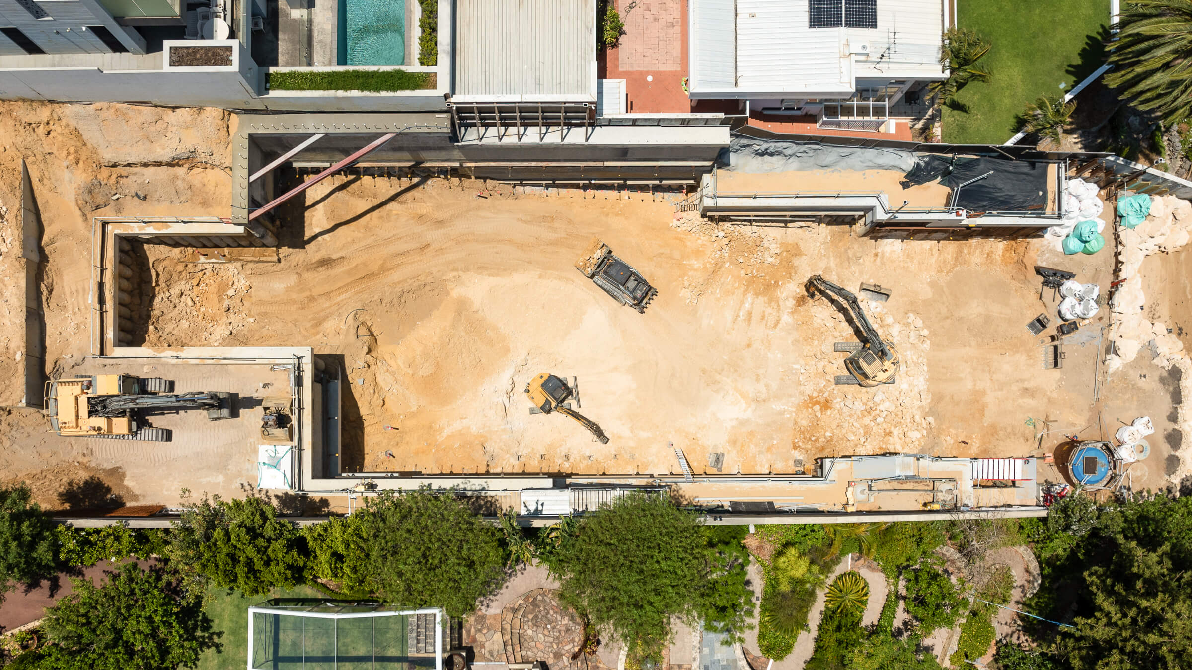 Aerial view of a construction site with three excavators working on leveled dirt surrounded by retaining walls and neighboring buildings.