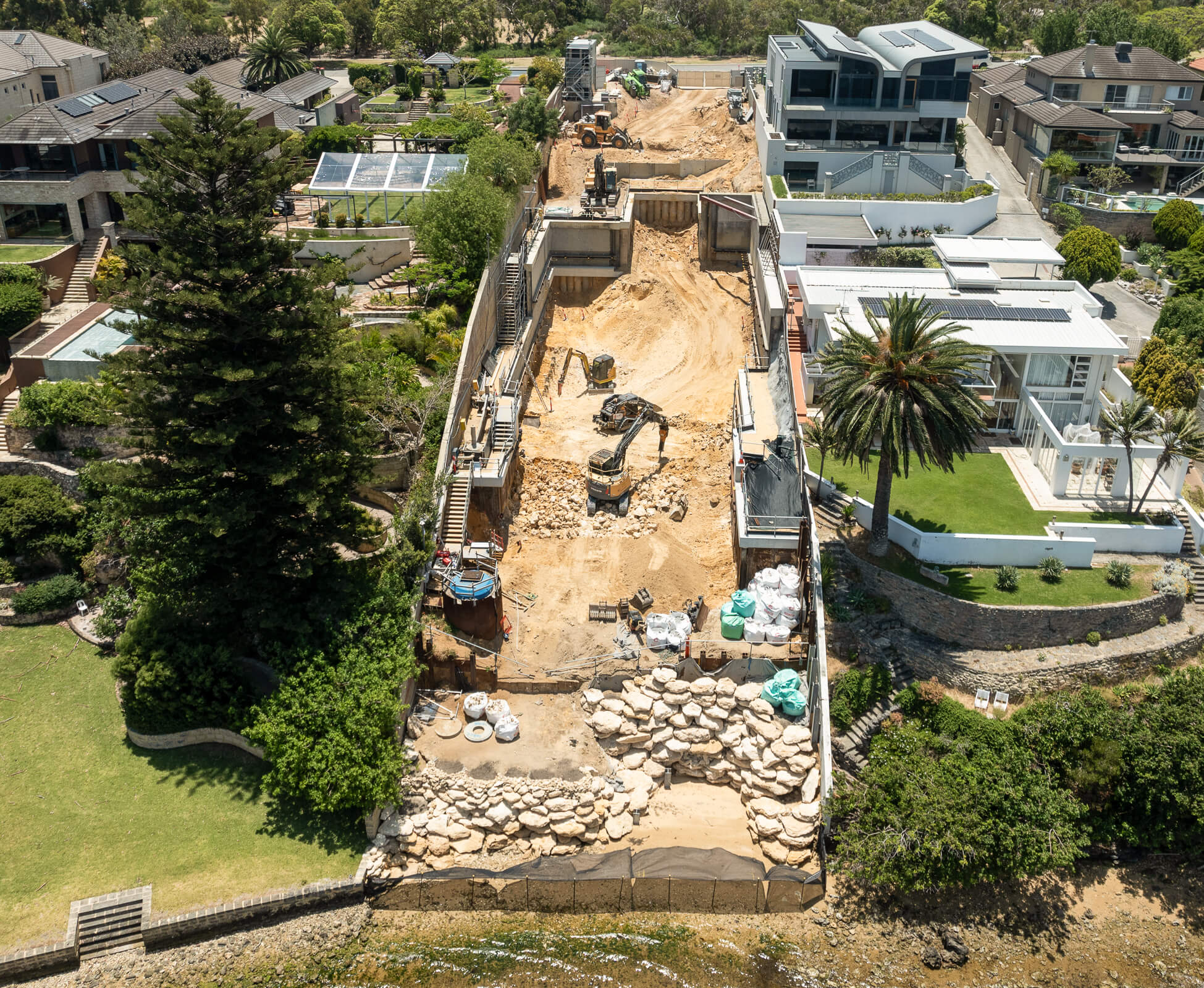 Aerial view of a large construction site between residential houses with excavators and stone retaining walls near a waterfront.