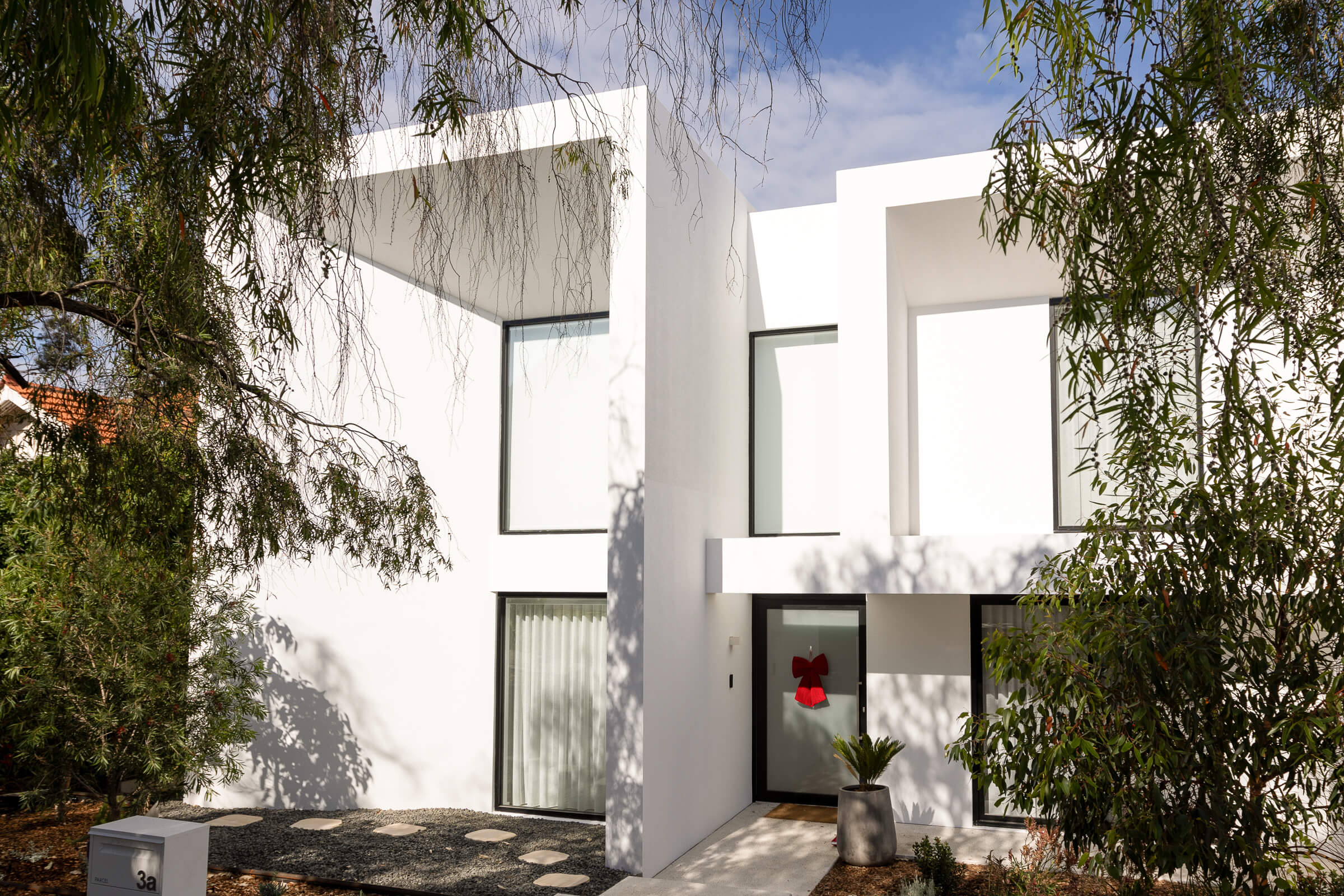 Modern white two-story house with large windows, a red bow on the front door, surrounded by trees.