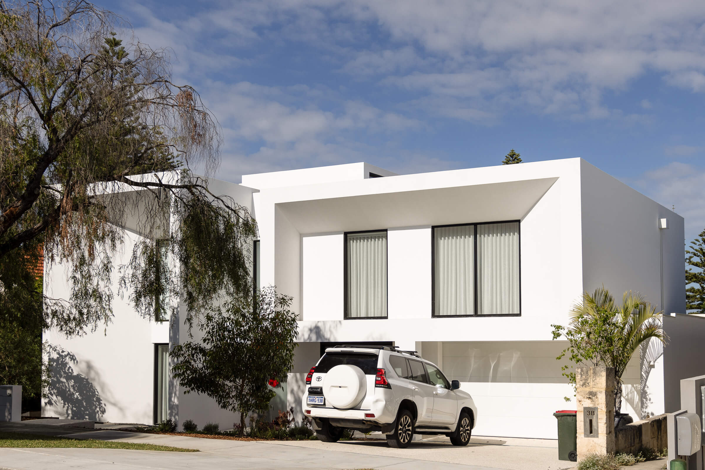 Modern white two-story house with large windows, a white SUV parked in the driveway, and surrounding trees under a partly cloudy sky.