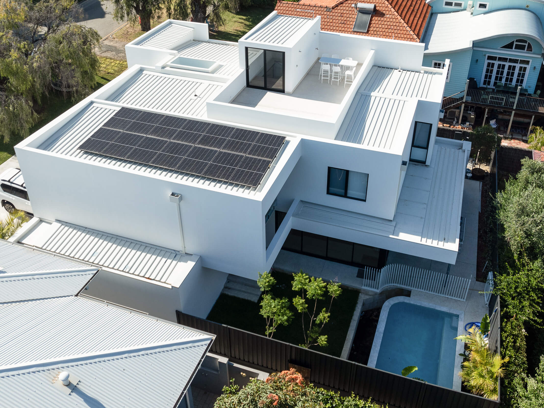 Aerial view of a modern white house with a flat roof featuring solar panels, a small rooftop patio with table and chairs, a backyard pool, and surrounding greenery.