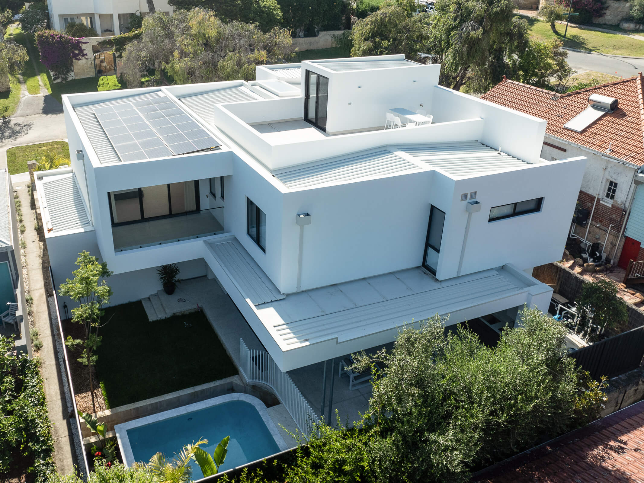 Modern white two-story house with solar panels on the roof, a small pool, and outdoor patio seating surrounded by greenery.