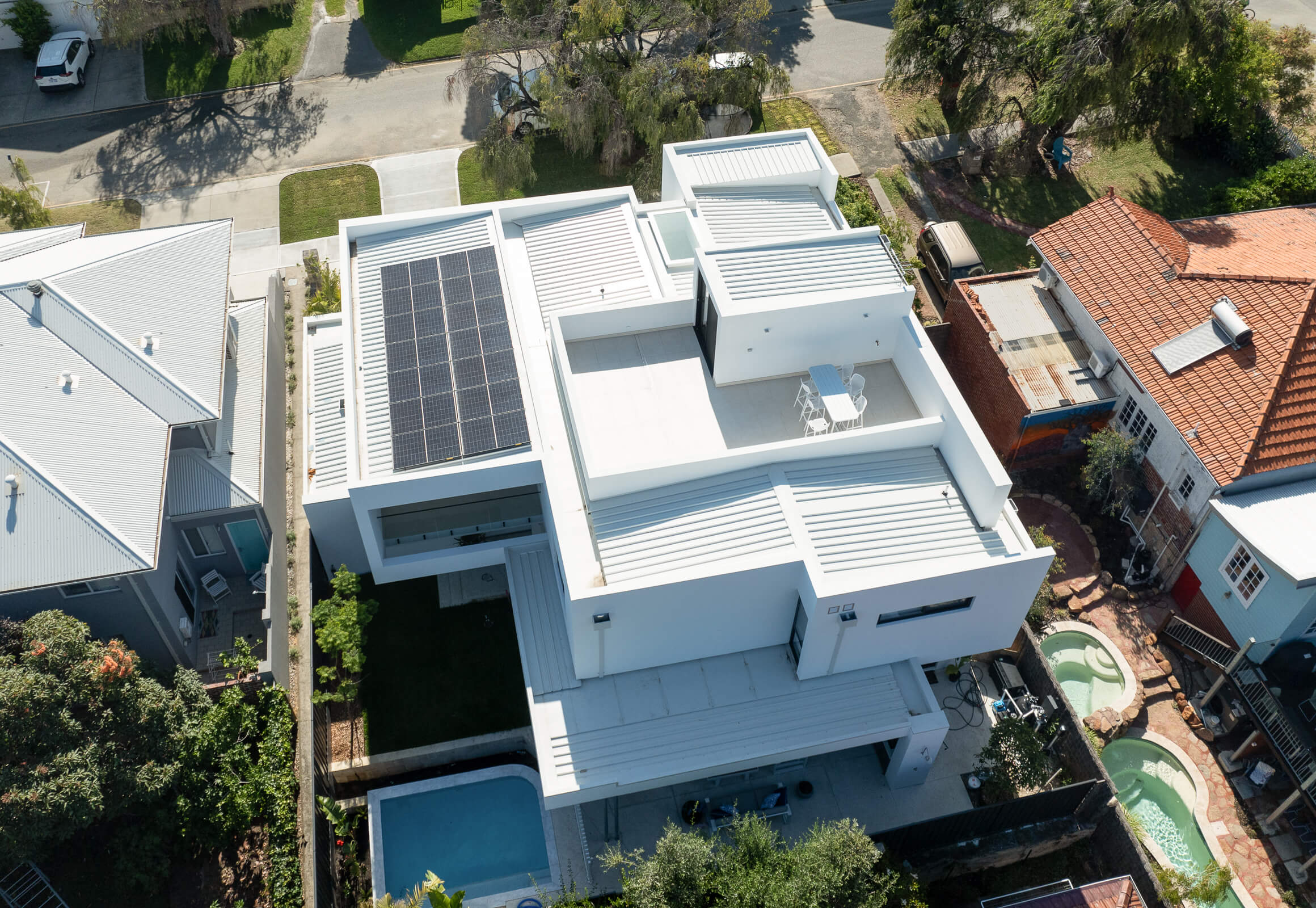 Aerial view of a modern white flat-roofed house with solar panels, rooftop terrace, and swimming pool in a suburban neighborhood.
