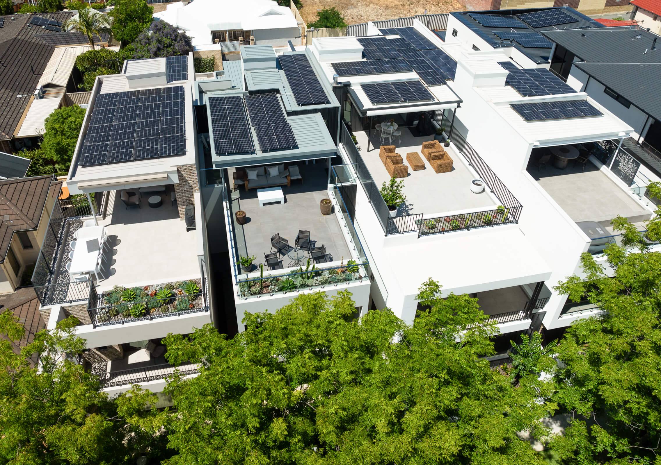 Modern white residential buildings with rooftop patios, solar panels, outdoor furniture, and surrounding green trees.