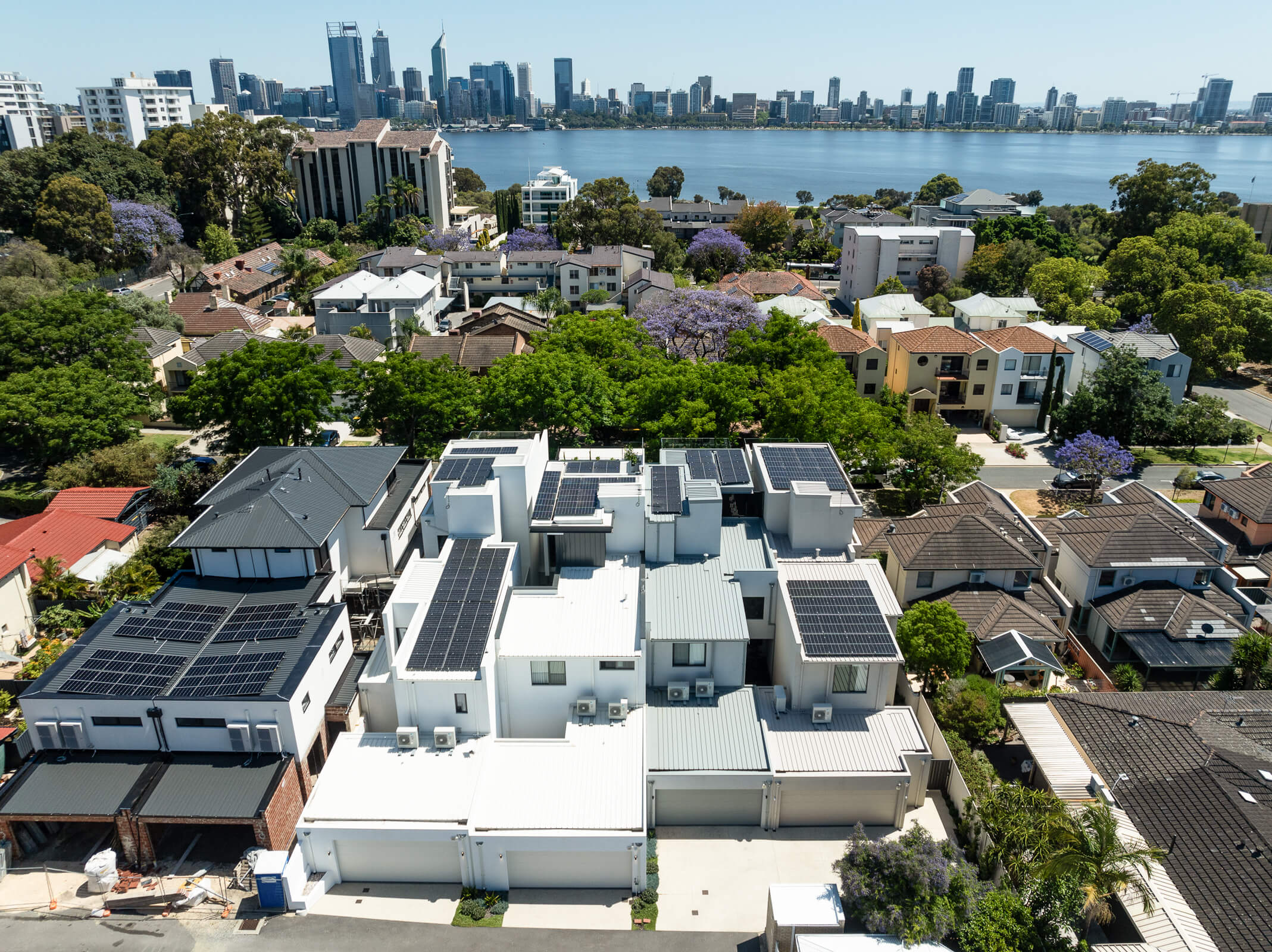 Aerial view of a residential neighborhood with modern houses featuring solar panels on rooftops, with a river and city skyline in the background.