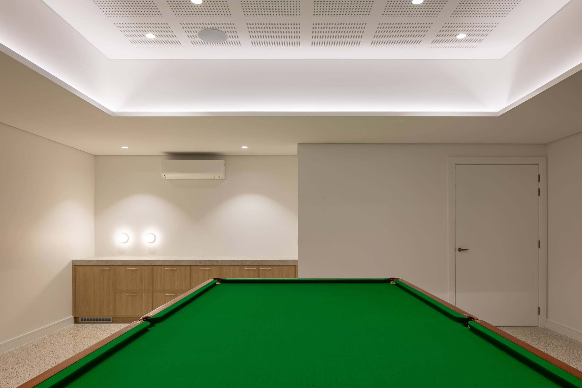 Minimalist indoor room with a green pool table, wooden cabinets, air conditioner, and closed white door.