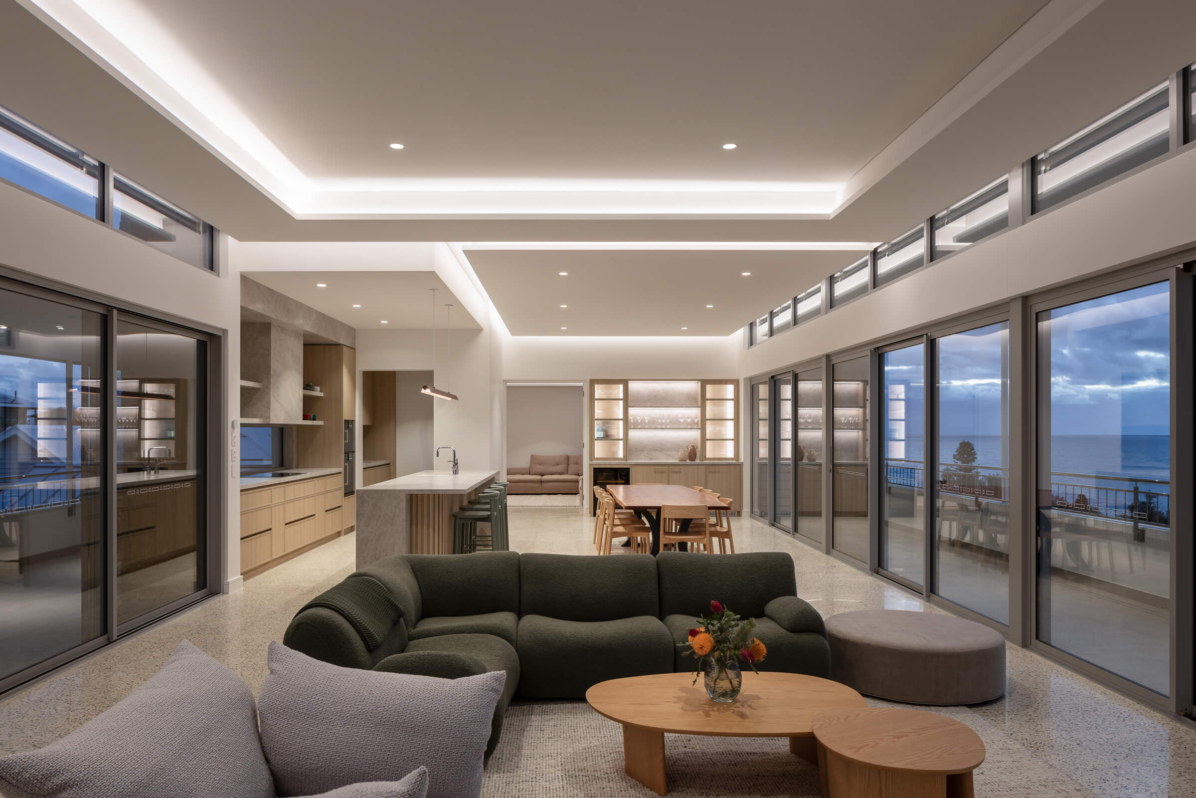 Modern open-plan living space with green sectional sofa, wooden coffee tables with flowers, dining table, kitchen island, and large windows overlooking ocean views at dusk.