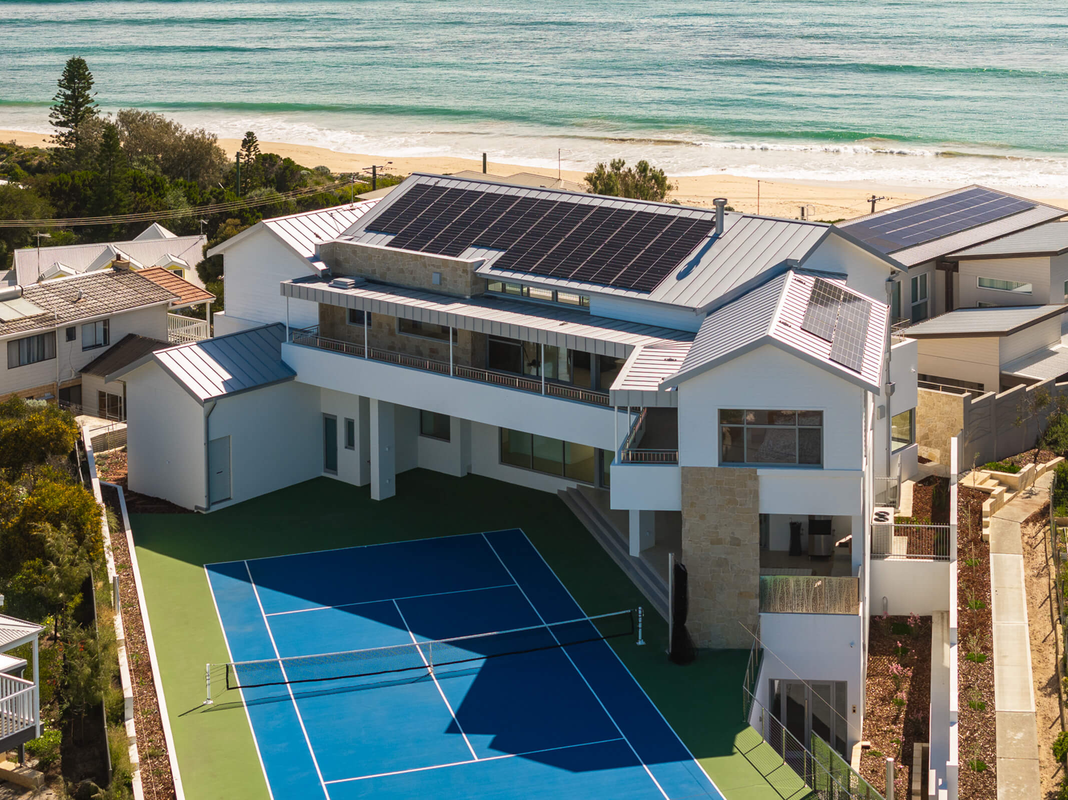 Modern beachfront house with solar panels on the roof and a blue tennis court in the backyard.