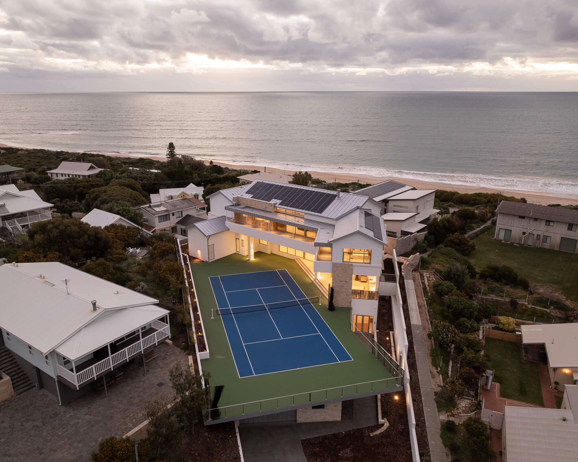 Aerial view of a modern coastal house with a rooftop tennis court, solar panels, and a beach in the background at dusk.
