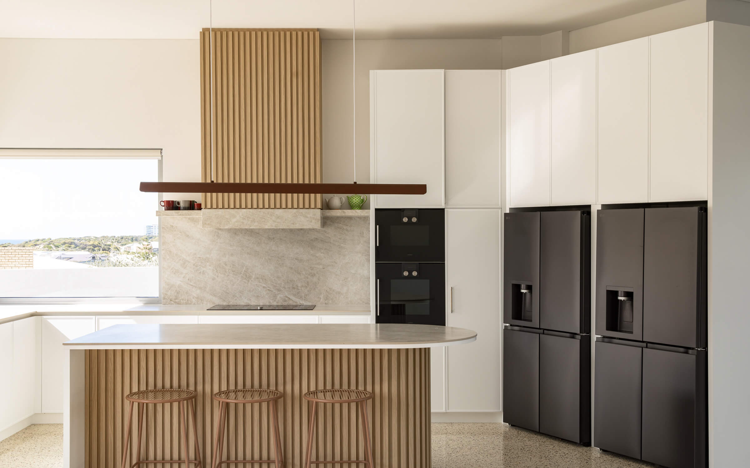 Modern kitchen with a marble island, three wooden stools, two black refrigerators, white cabinets, and a panoramic window.
