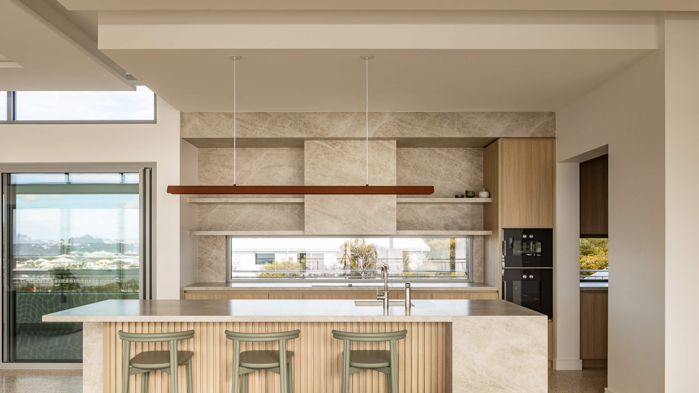 Modern kitchen with beige marble countertops, wooden cabinetry, three light green bar stools, and large window.