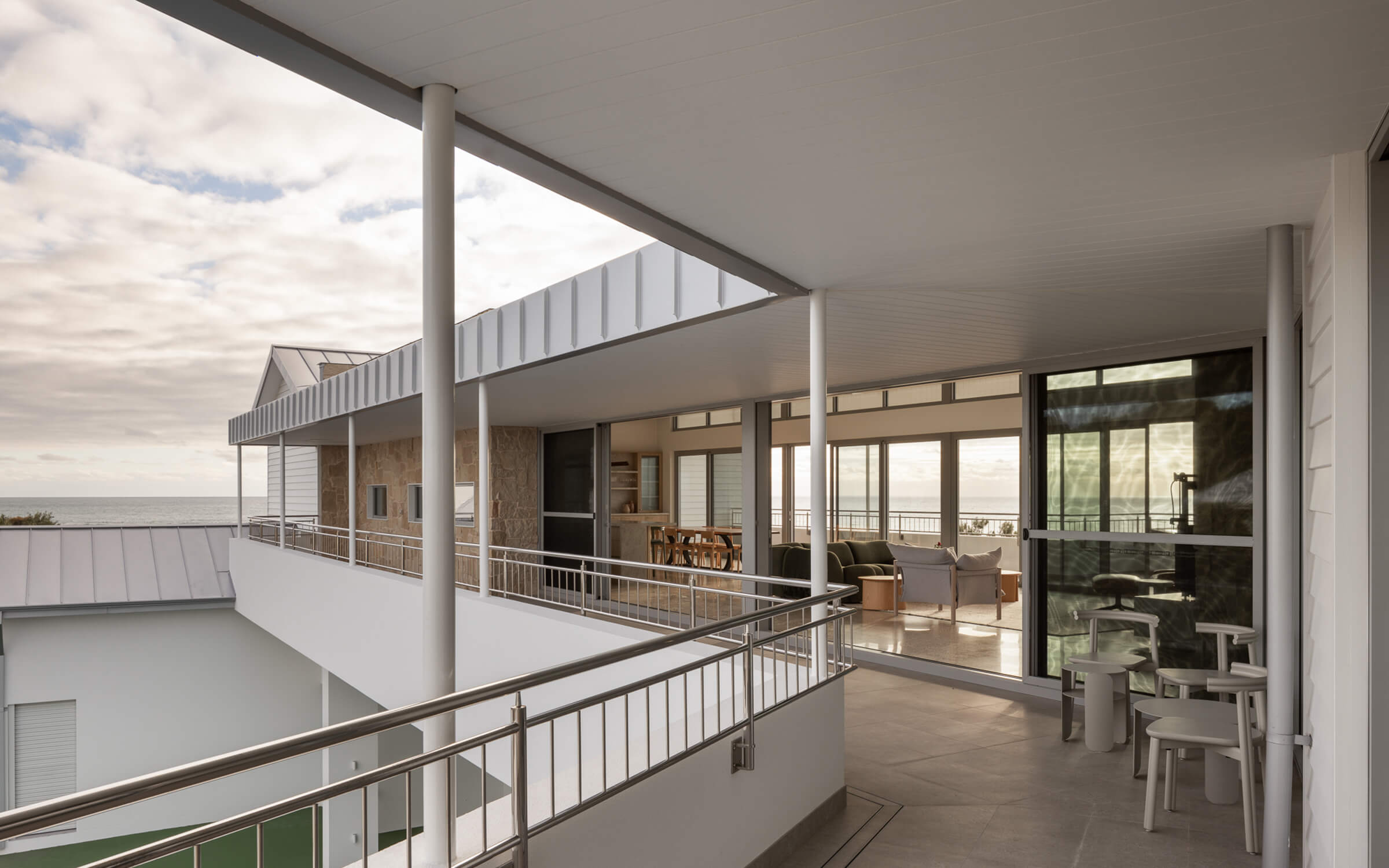 Modern open-air balcony with metal railings overlooking ocean, adjacent to living room with large windows.