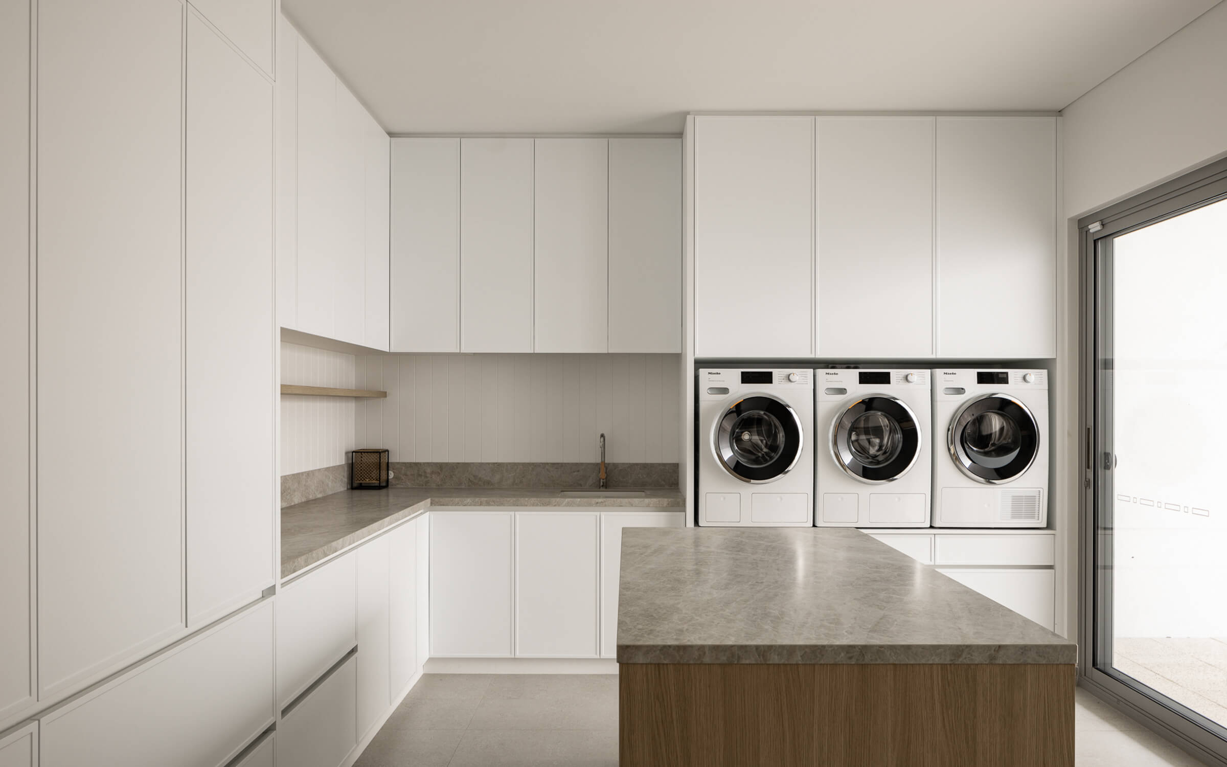 Bright modern laundry room with white cabinets, three front-loading washing machines, a gray countertop island, and large sliding glass door.