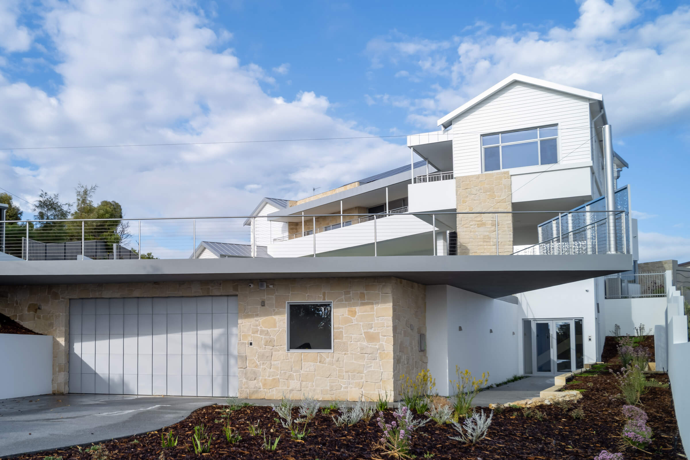 Modern multi-level house with stone and white siding exterior, metal railings, and landscaped front yard under a partly cloudy sky.
