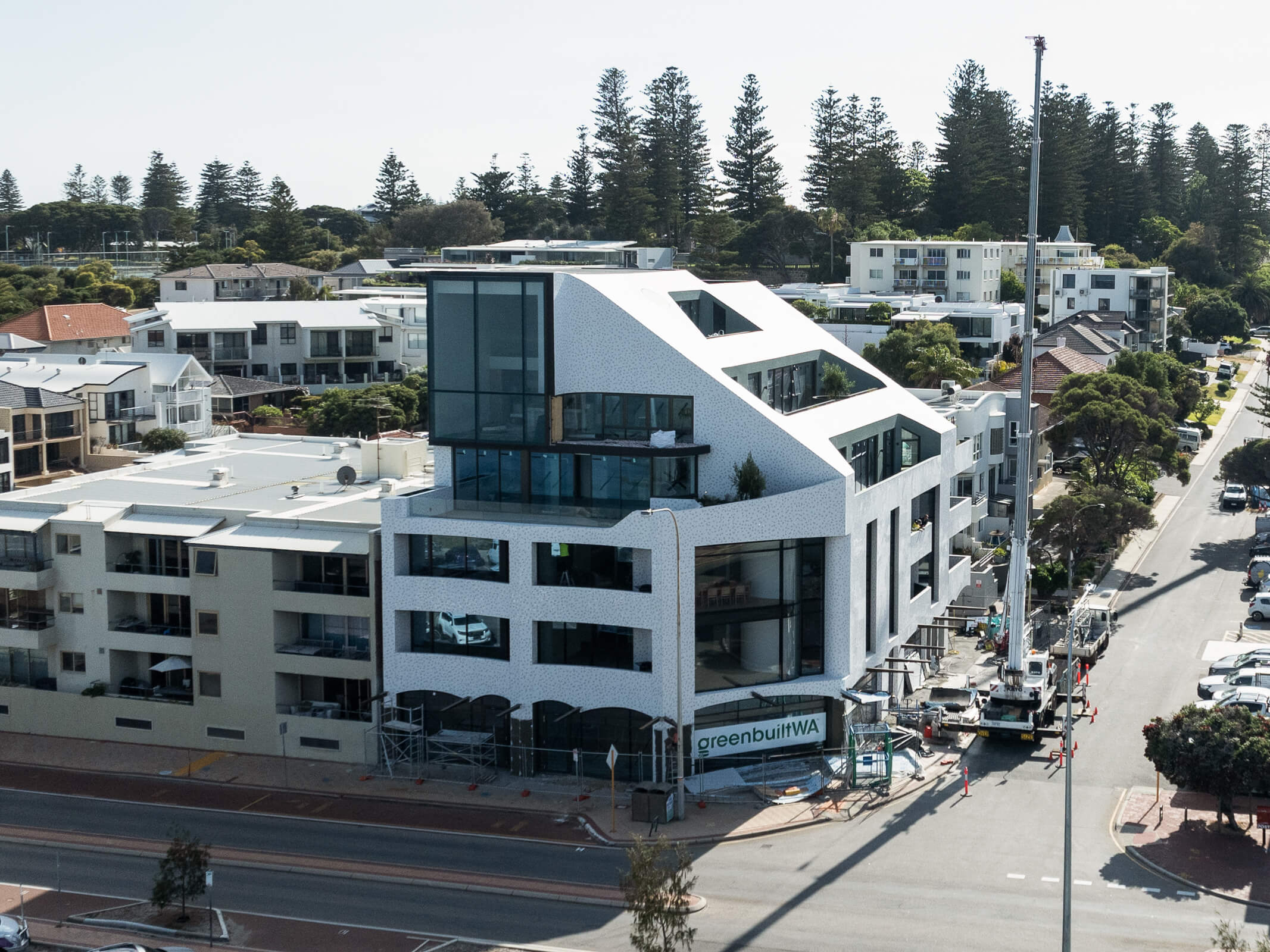 Modern white multi-story building with large windows under construction in a suburban area lined with trees and residential buildings.