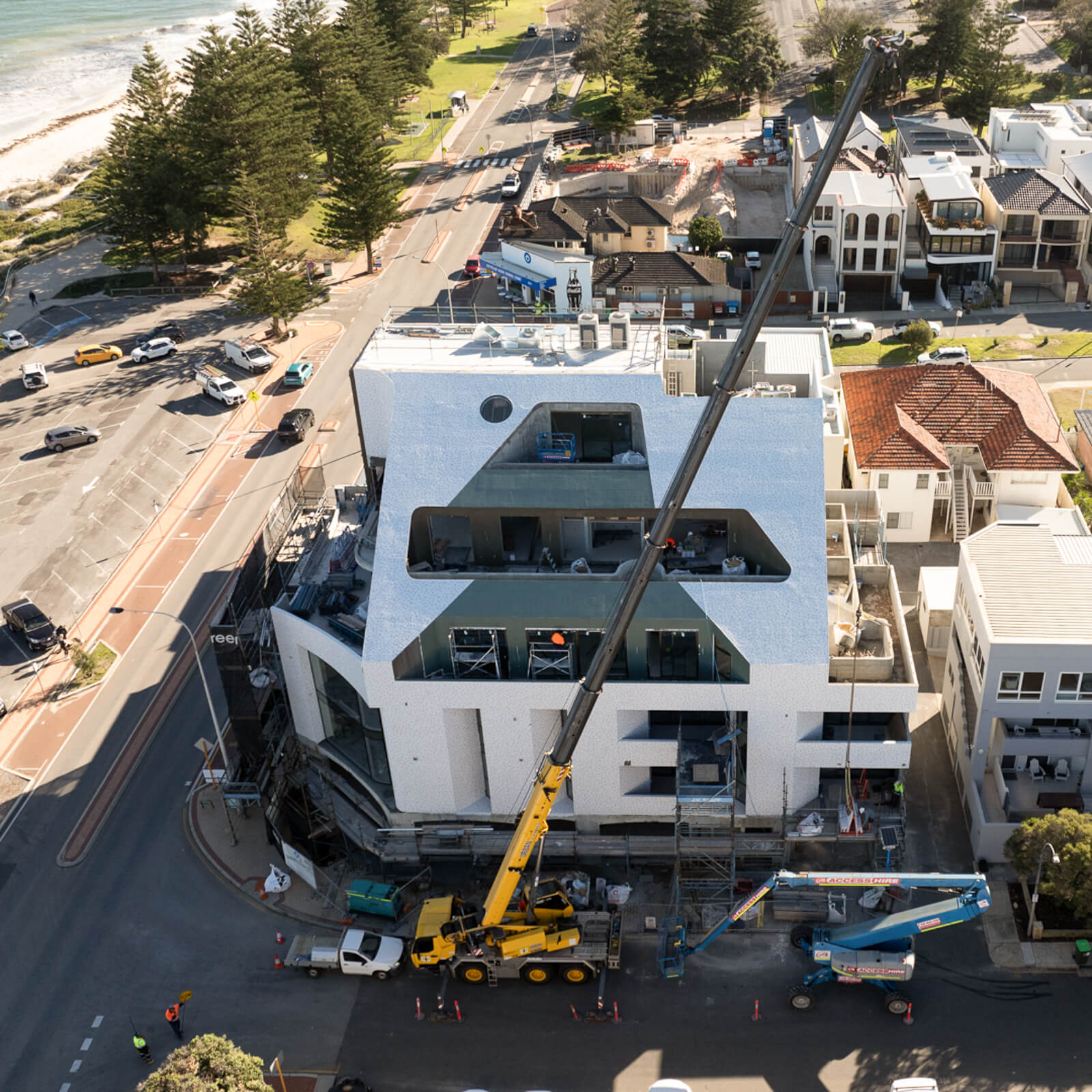 Aerial view of a modern building under construction near the seaside with a large yellow crane and surrounding roads.